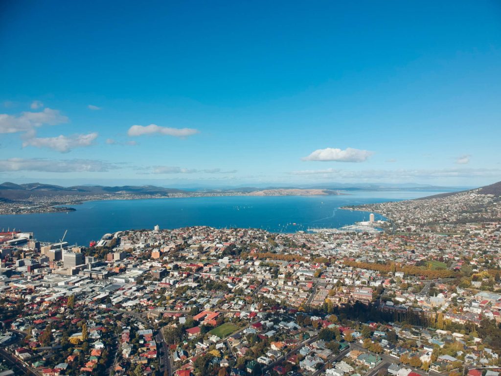 The photograph is an aerial shot of Hobart, Tasmania, one of Drive Car Hire's main locations. There are houses and neighbourhoods and beautiful blue open waters and hilly terrain in the background.