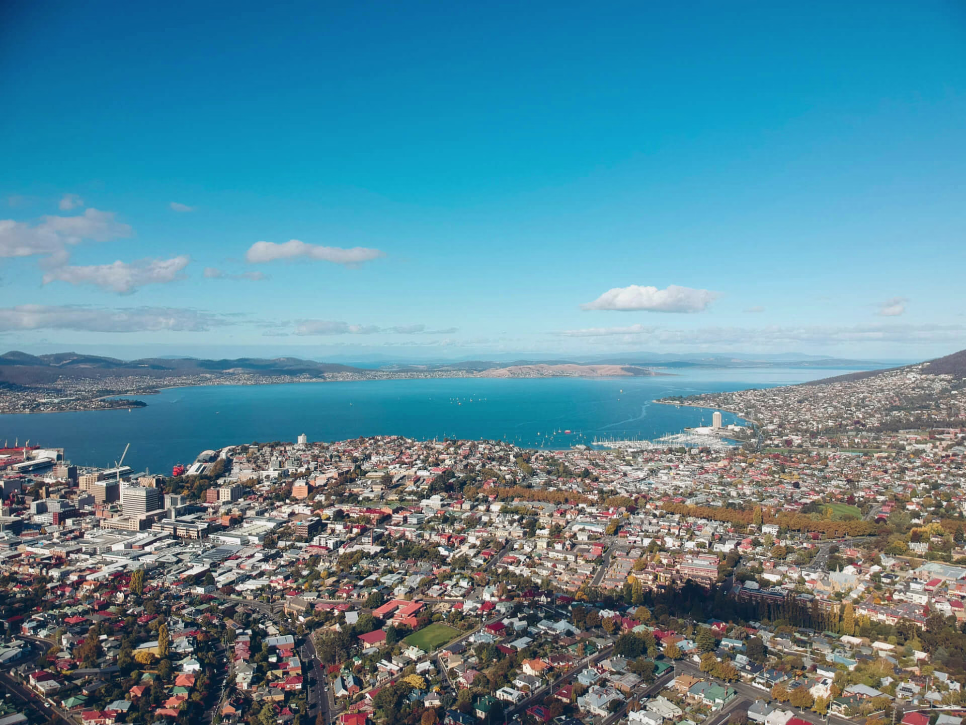 The image depicts a stunning, nearly cloudless view of Hobart city, taken from above. Vast turquoise waters, sweet neighbourhoods, and an expanse of sky make for a lovely photo! Book your car with Drive Car Hire Tasmania and explore Hobart today.