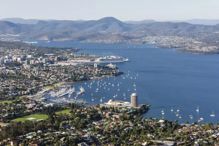 The picture is an aerial shot of Hobart. Depicted is the casino, the harbour, some sports fields, lots of scattered, docked boats, a bridge, a few neighbourhoods, and mountains in the background. 