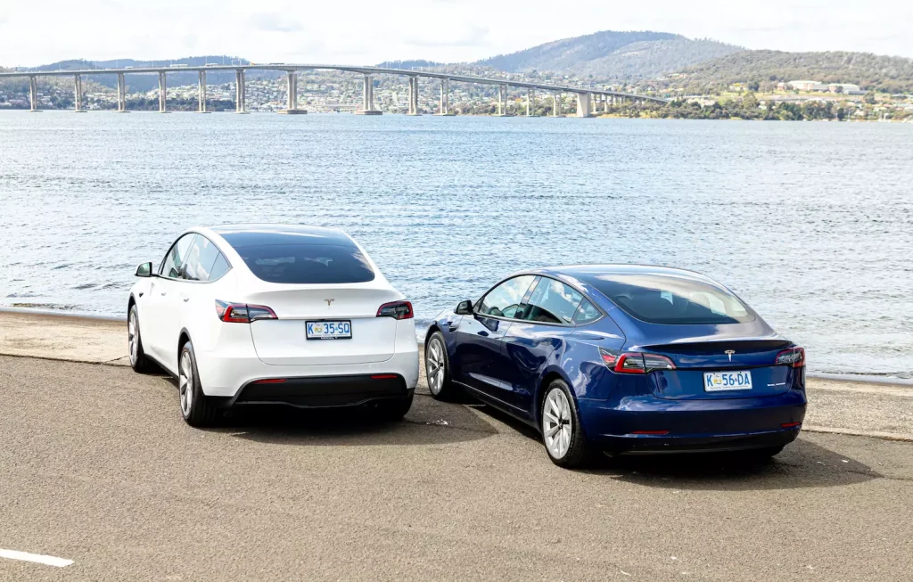 Two Teslas - one white and one blue - look out at the water and a bridge in Hobart. 