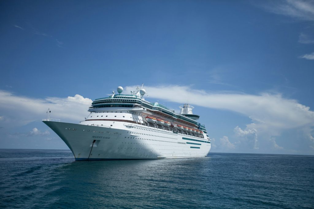 The photograph contains a cruise ship on the open water on a sunny, bright day. Lots of blue hues and a few wispy clouds circle the ship.