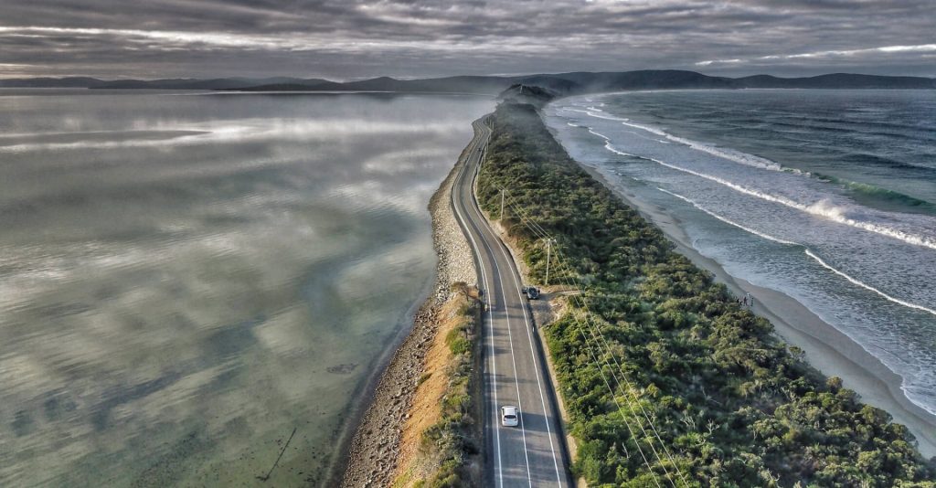 This is a moody shot of Bruny Island - sea on two sides of a thinner road. A white car travels from the bottom upward in the picture towards the mountains.