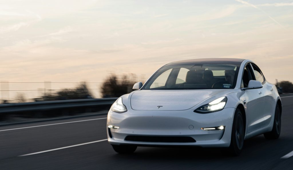 This picture shows a Tesla on the road, traveling quite fast, as the background is blurred. A pastel sky behind the electric vehicle alludes to an early morning or sunset drive...