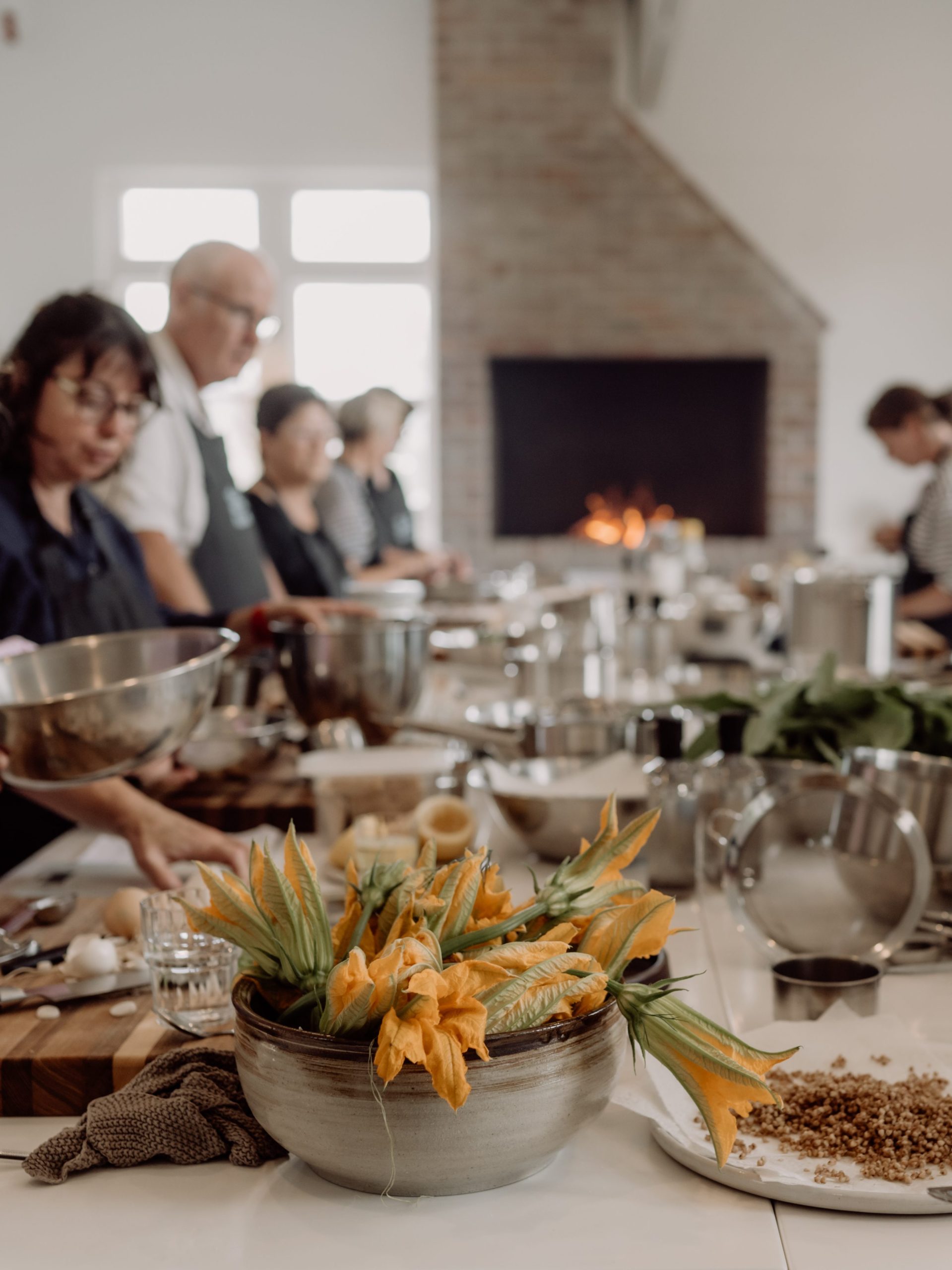 This is a picture of a Guest Chef cooking masterclass at The Agrarian Kitchen in New Norfolk. Beautiful ingredients, flowers, and grains are the focus of the shot and we see students hard at work in the background. 
Shot by Anna Critchley.