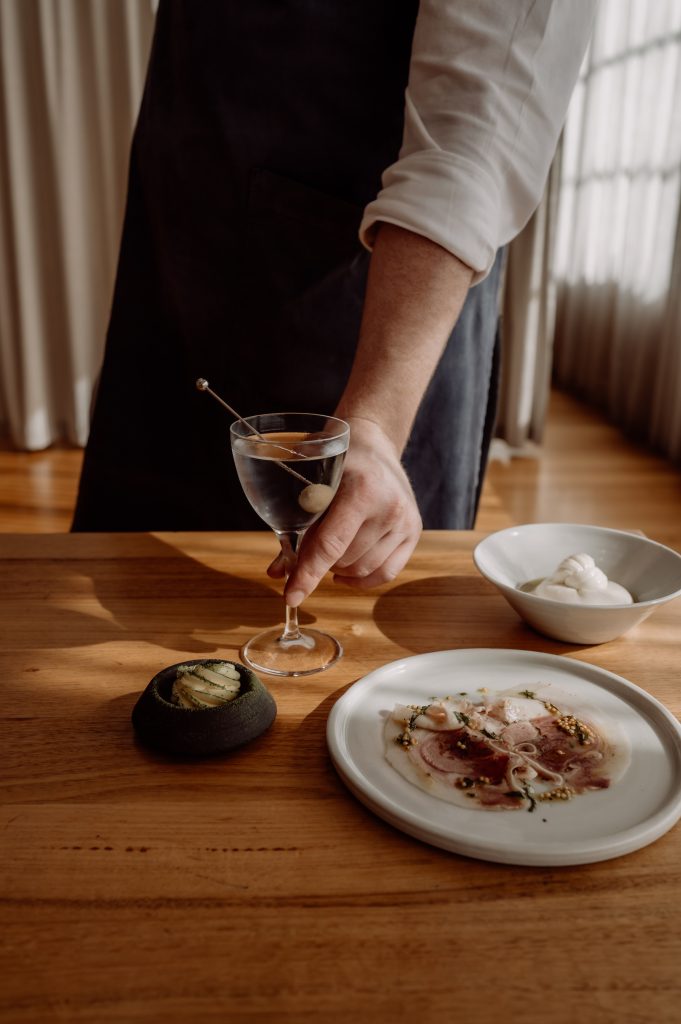 Photography by Anna Critchley. A waiter serves a beautiful martini alongside some thinly sliced meat, homemade butter, and ricotta at The Agrarian Kitchen.
