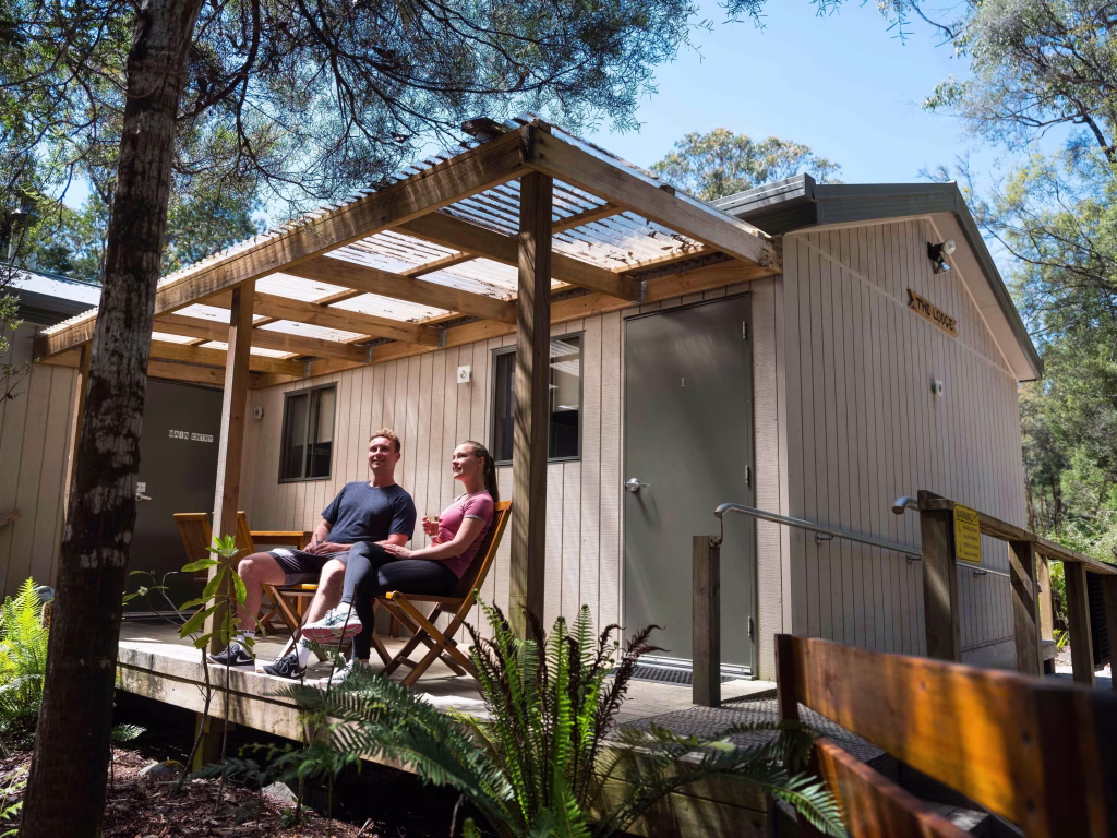 Photo credit: Tahune Adventures Tasmania. A man and woman sit outside on wooden chairs gazing out from their porch at one of Tahune's accommodations.