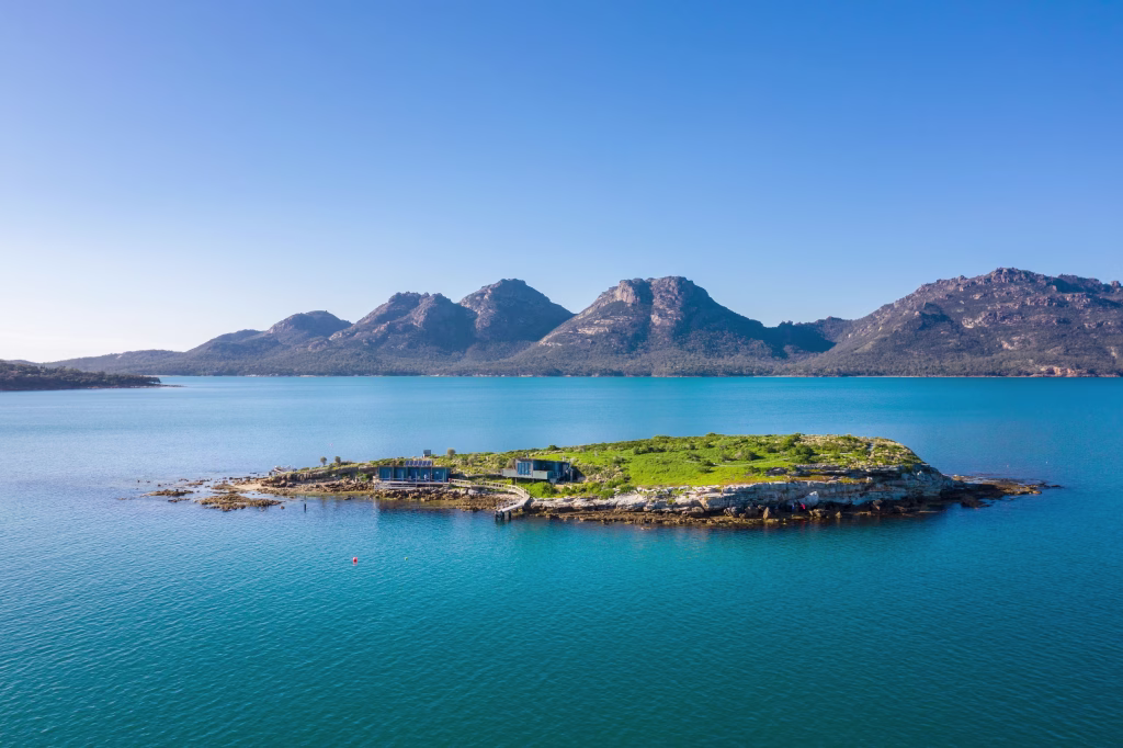 Taken by Luke Tscharke, this photo depicts Picnic Island in Tasmania with clear, turquoise waters, greenery, and the Hazards mountain range as a backdrop. It's a bluebird day without a cloud in the sky.