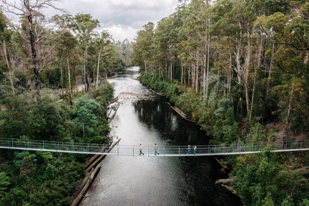 The image, taken by Tourism Australia, depicts the Swinging Bridges Walk at Tahune Adventures. Two people walk across an elevated hanging bridge over a river, framed by tall trees and greenery on both sides.