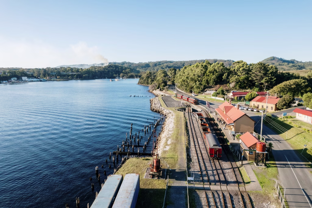An aerial shot of the West Coast Wilderness Railway train at one of the stations next to clear, blue water. Photograph by Tourism Australia