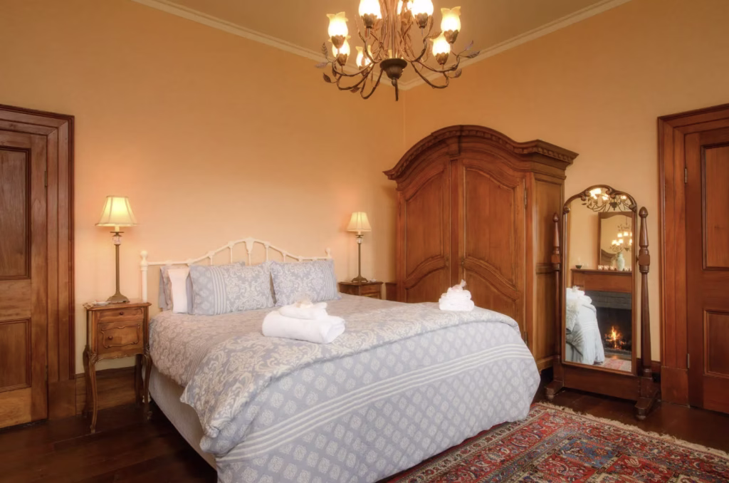 A light blue made bed with fresh towels in a historic bedroom with a fireplace, armoire, and mirror at the Rathmore House.