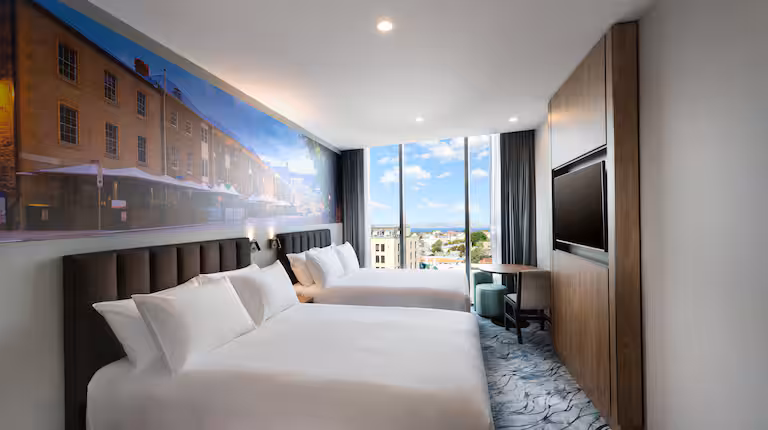 A room at the DoubleTree by Hilton Hobart with two queen beds made up with crisp white linens. The room overlooks the Hobart CBD and we can see the water in the distance. The backsplash features the Salamanca restaurant strip.