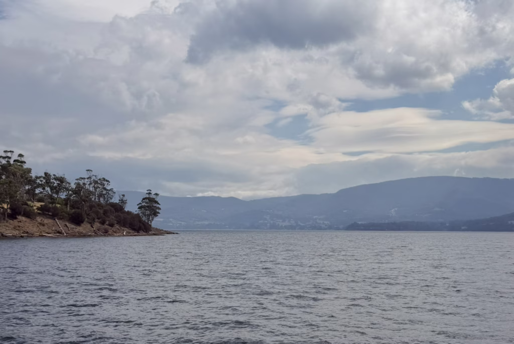 A shot from the ferry on the way to Bruny Island. Water in the foreground, some land on the left side, and mountainous and hilly Tasmanian backdrop. It's a cloudy, greyish day.