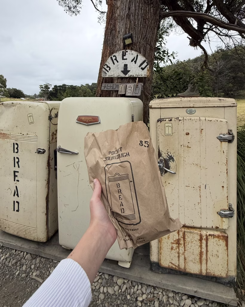 A hand holds a "Pocket sourdough" bread from the Bruny Baker, a roadside bakery with several bread fridges in the background.