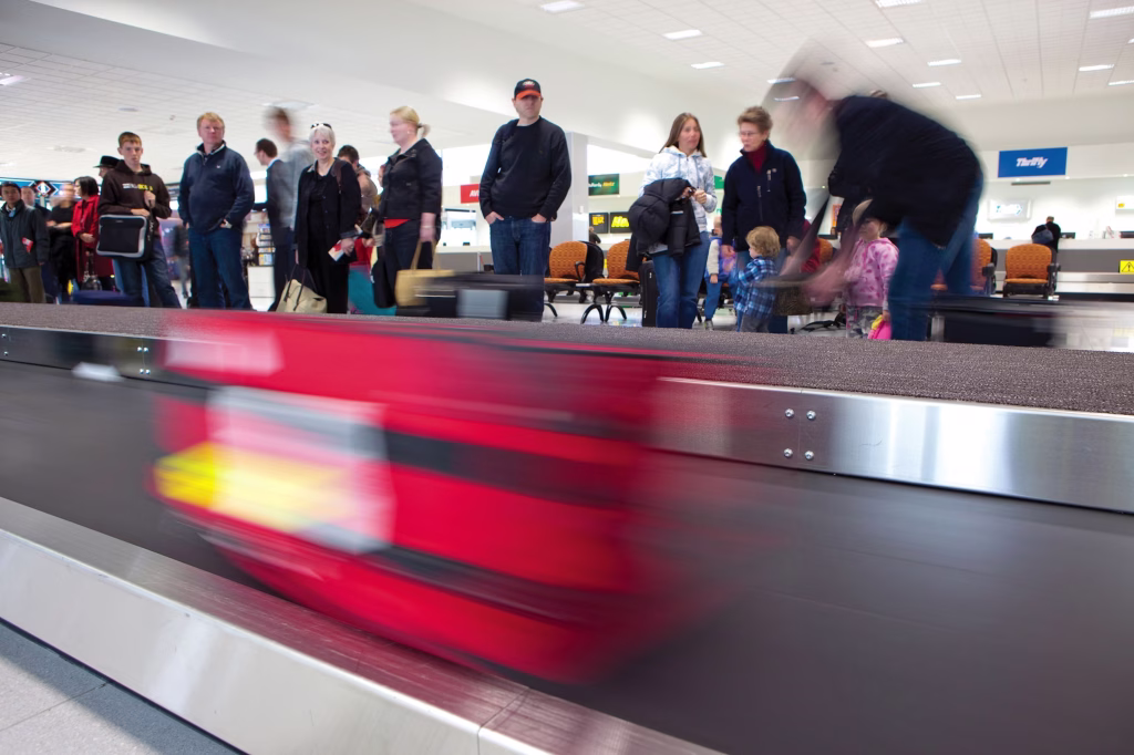 A luggage bag in red zips by on a bag return at an airport in Tasmania, Australia. Behind the baggage claim, people congregate, waiting for their items.

Photo by Rob Burnett.
