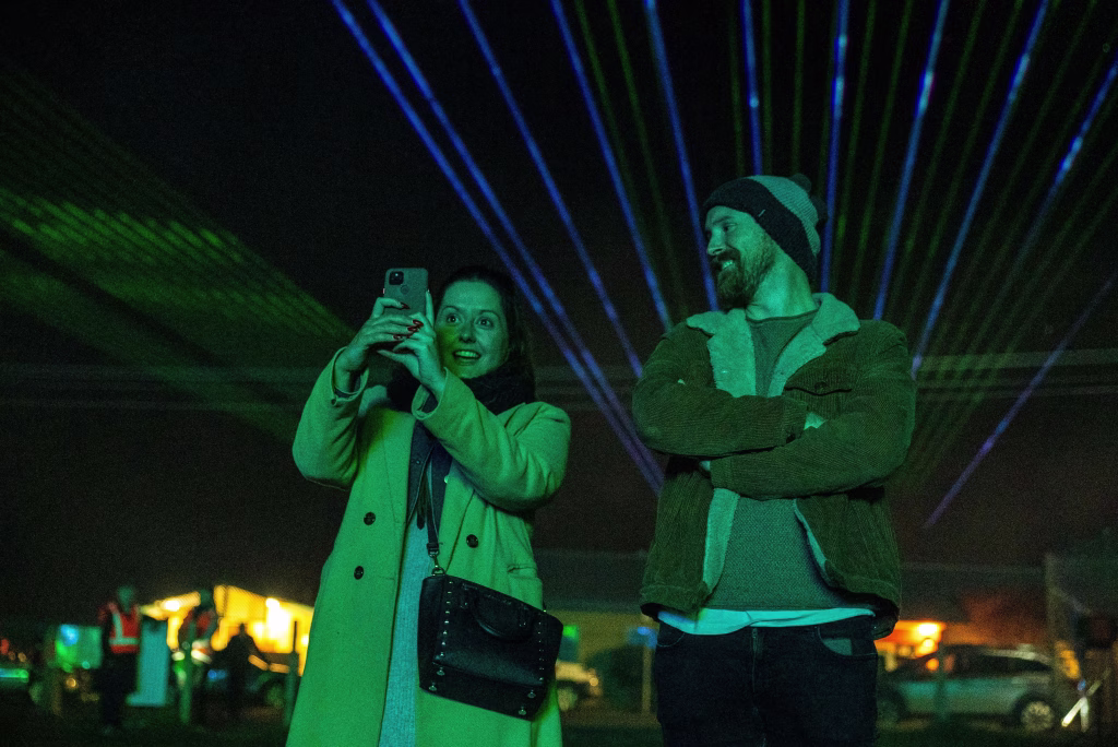 A woman takes a picture with her phone of the Bicheno Beams laser light show as a man smiles at her with his arms crossed. Photo: Adam Reibel